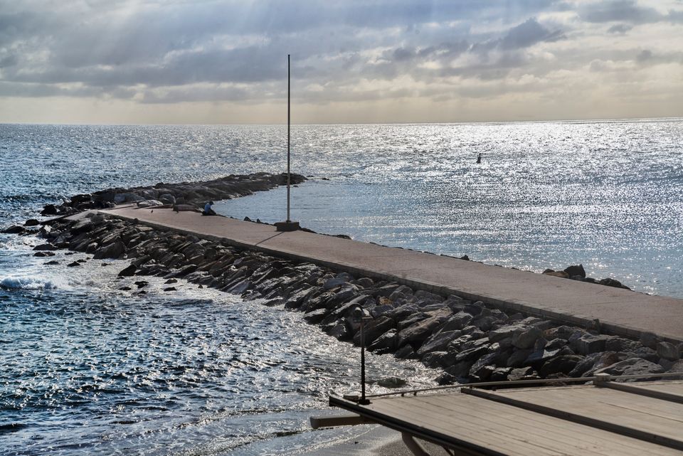 Scena di spiaggia calma con una diga pavimentata che si estende nell'oceano sotto un cielo nuvoloso.