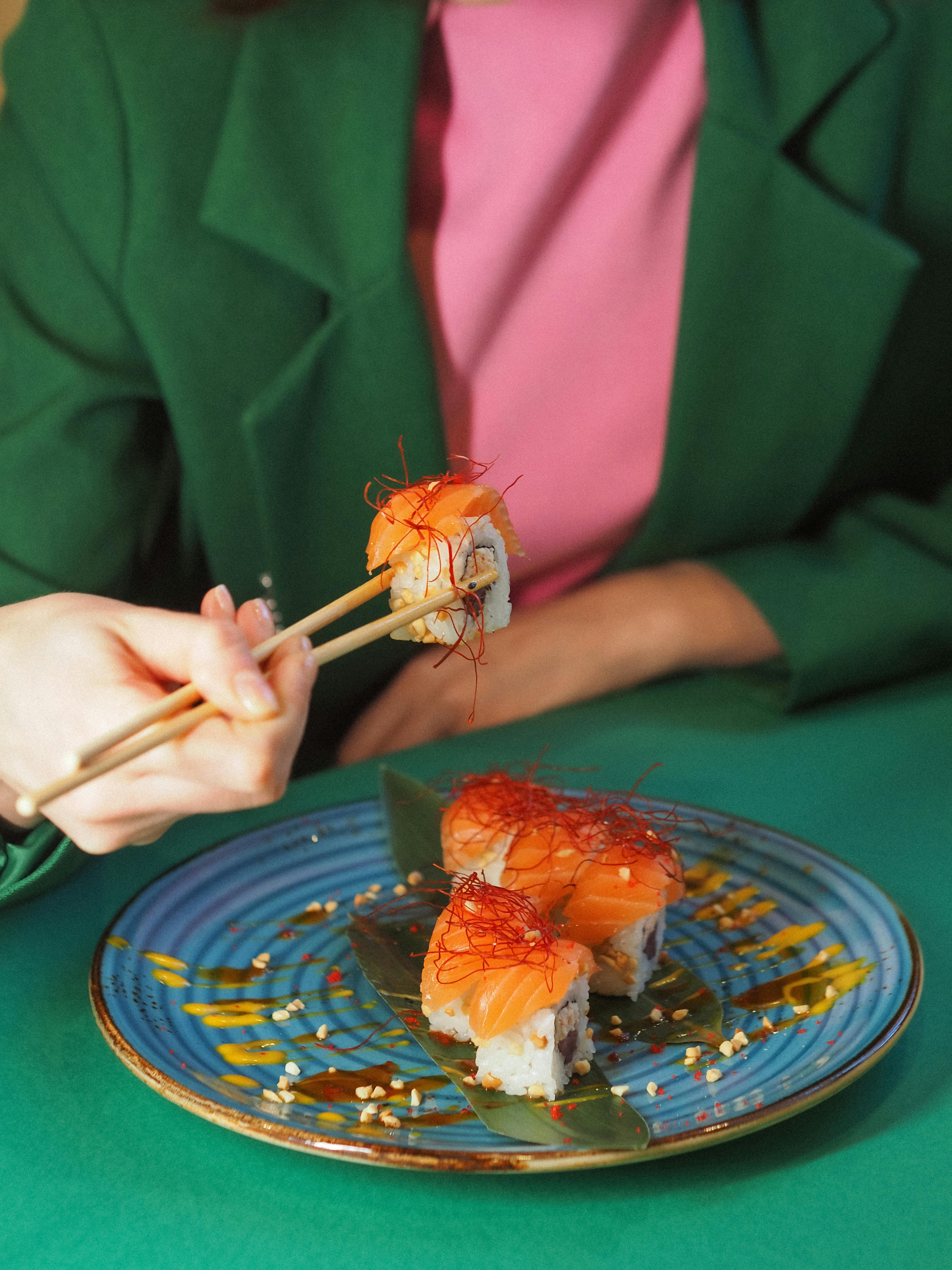 A person in a green jacket eating sushi with chopsticks on a colorful plate.