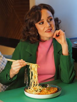 A woman in a green jacket and pink top dining on noodles at a restaurant table.