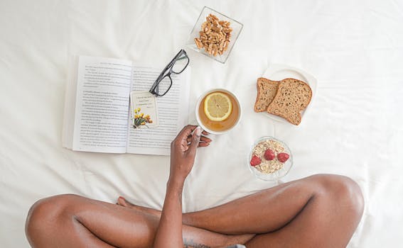 Person Holding White Ceramic Mug With Lemon Near Book and Sliced Bread on White Comforter