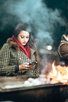 Woman in winter clothing using phone by open fire on a cold night.