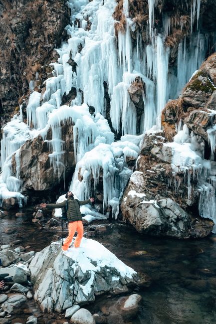 A person stands embracing nature at a frozen waterfall in Gulmarg, showcasing the icy beauty of winter.