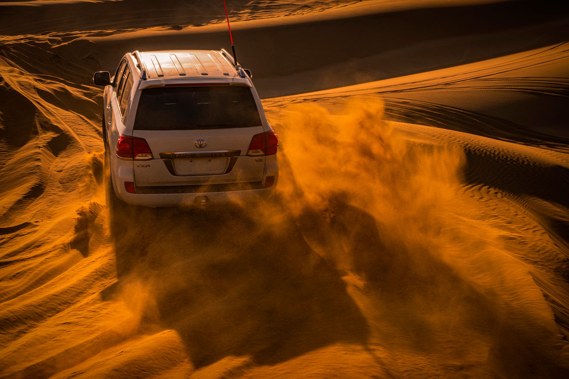 A Toyota Land Cruiser drives through sandy desert dunes, kicking up clouds of sand in its wake.