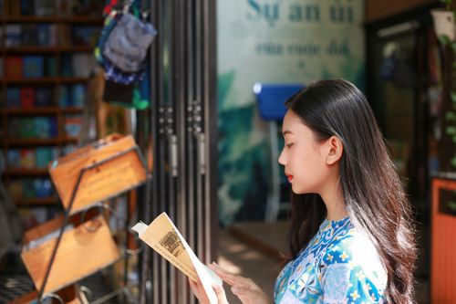 Free Side view of a young woman reading a book in a sunlit library with a blurred background. Stock Photo