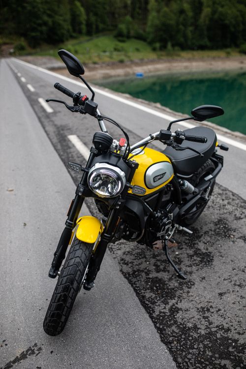 Free A vibrant yellow scrambler motorbike parked on a scenic roadside with lush greenery. Stock Photo