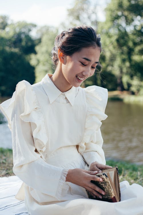 Smiling Young Woman in White Dress Holding Book in Park