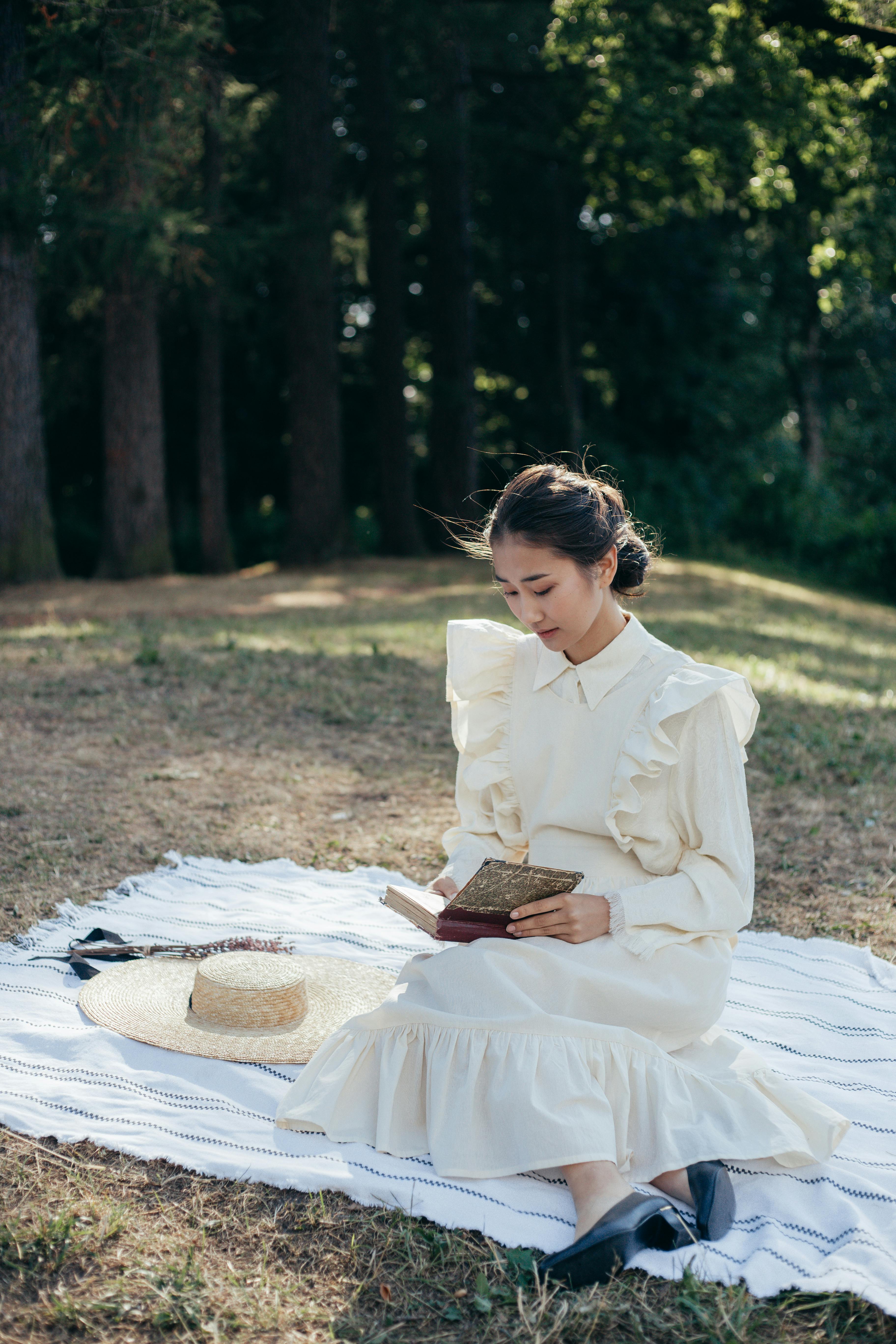 A young woman in a vintage dress reads peacefully on a picnic blanket in a tranquil park setting.
