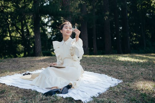 Young Woman in White Dress Sitting on Picnic Blanket in Park, Holding Dried Flowers