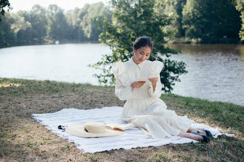 Free Young Woman in White Dress Reading Letter in Riverbank Stock Photo