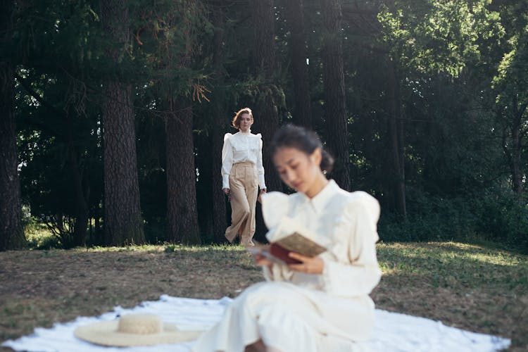 Young Woman In White Dress Reading Book On Picnic Blanket In Park