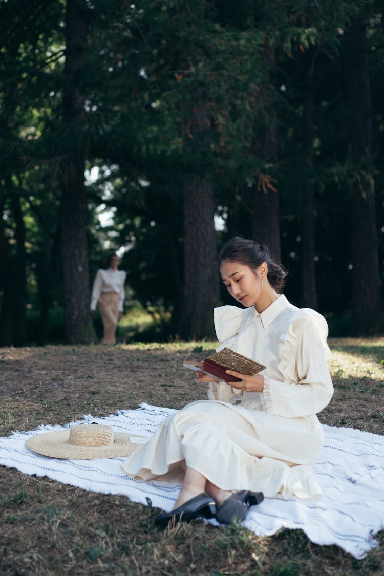 Young Woman In White Dress Reading Book On Picnic Blanket In Park