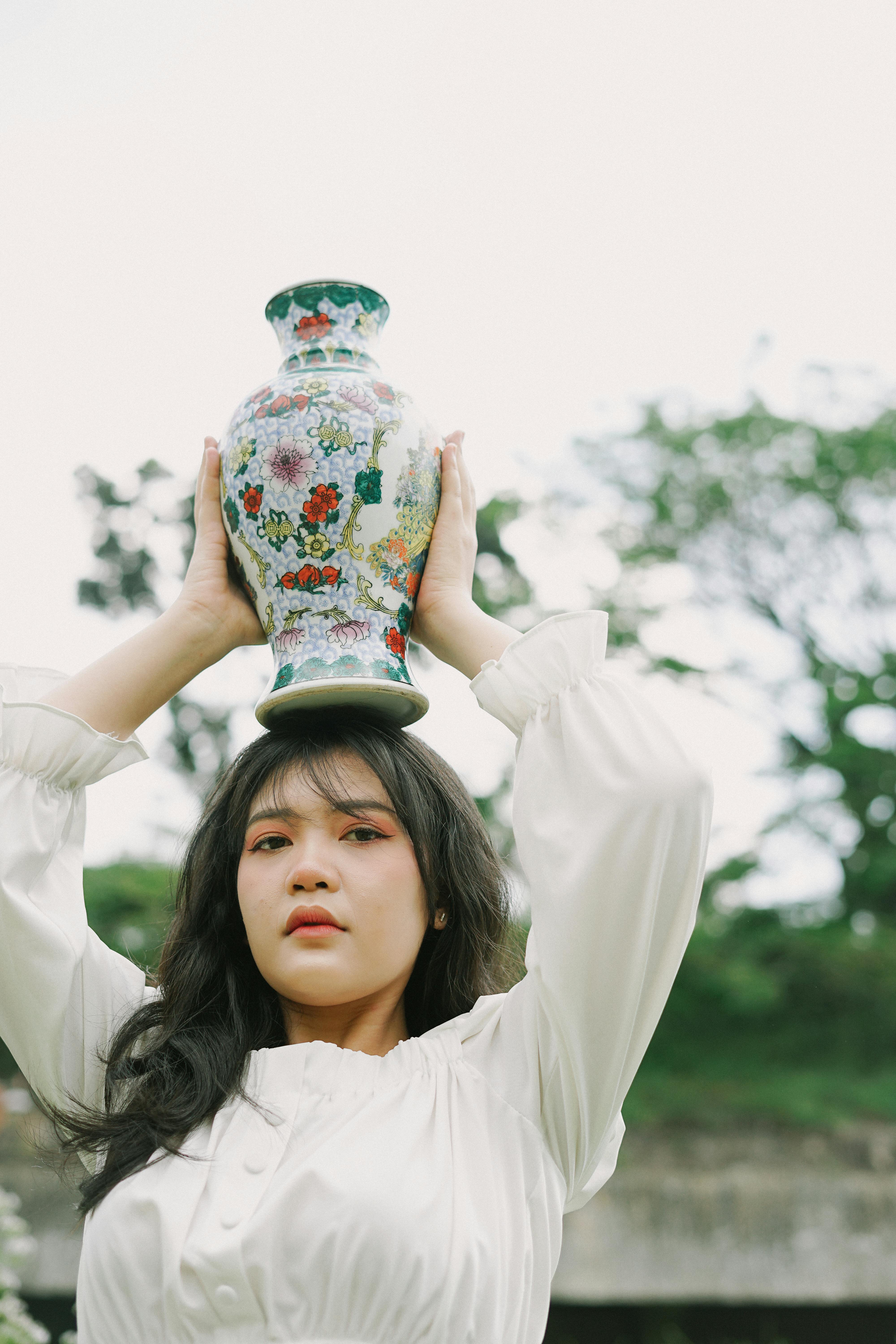 Portrait of a young woman holding a floral vase on her head in an outdoor setting.