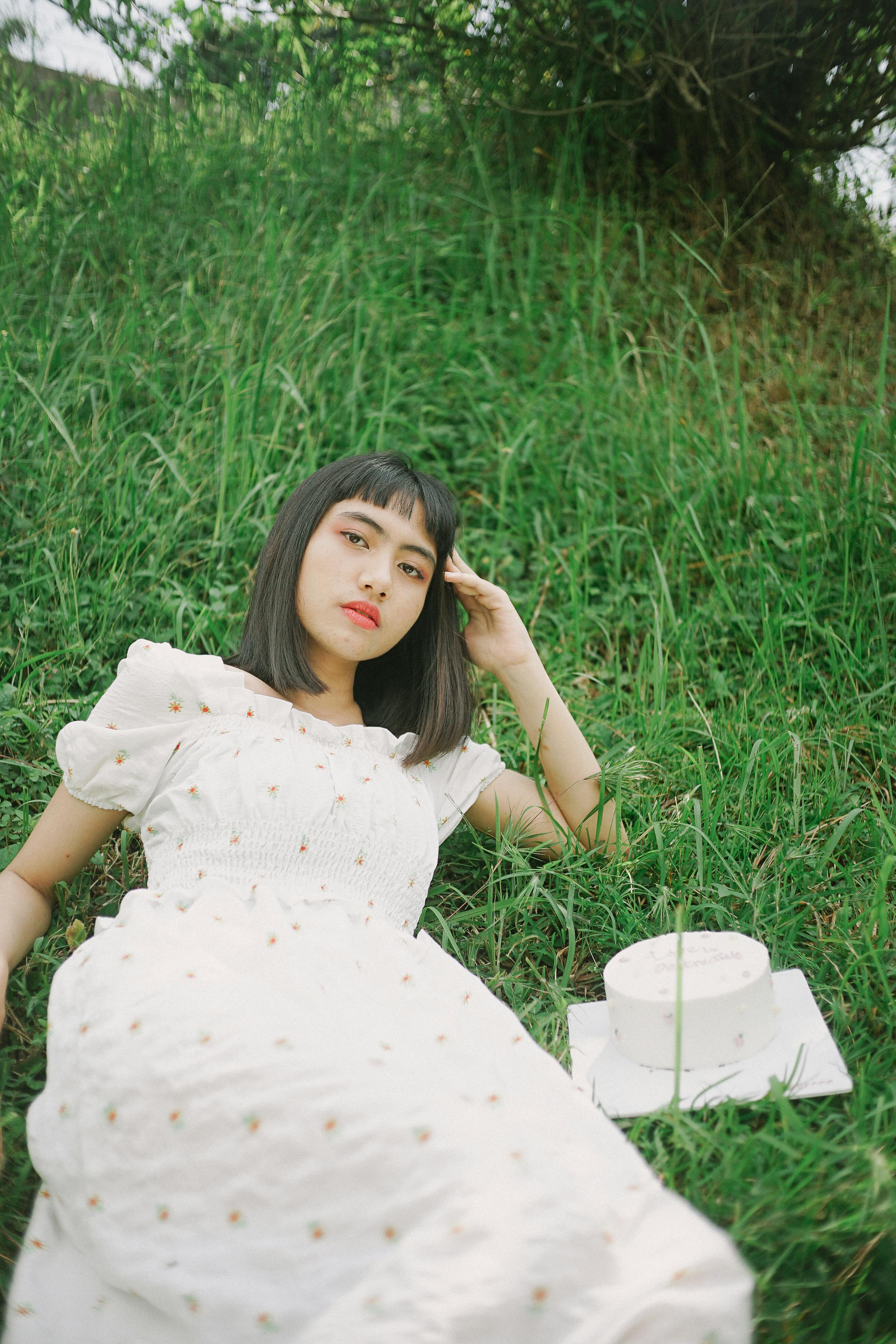 A young woman in a white dress lying on green grass beside a cake, exuding tranquility.