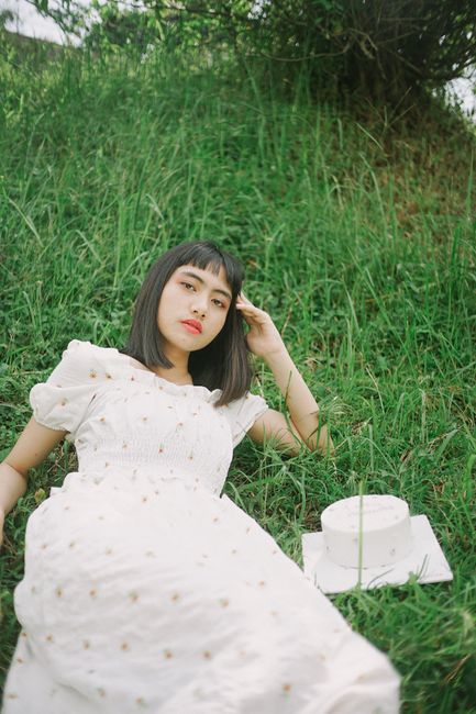 A young woman in a white dress lying on green grass beside a cake, exuding tranquility.