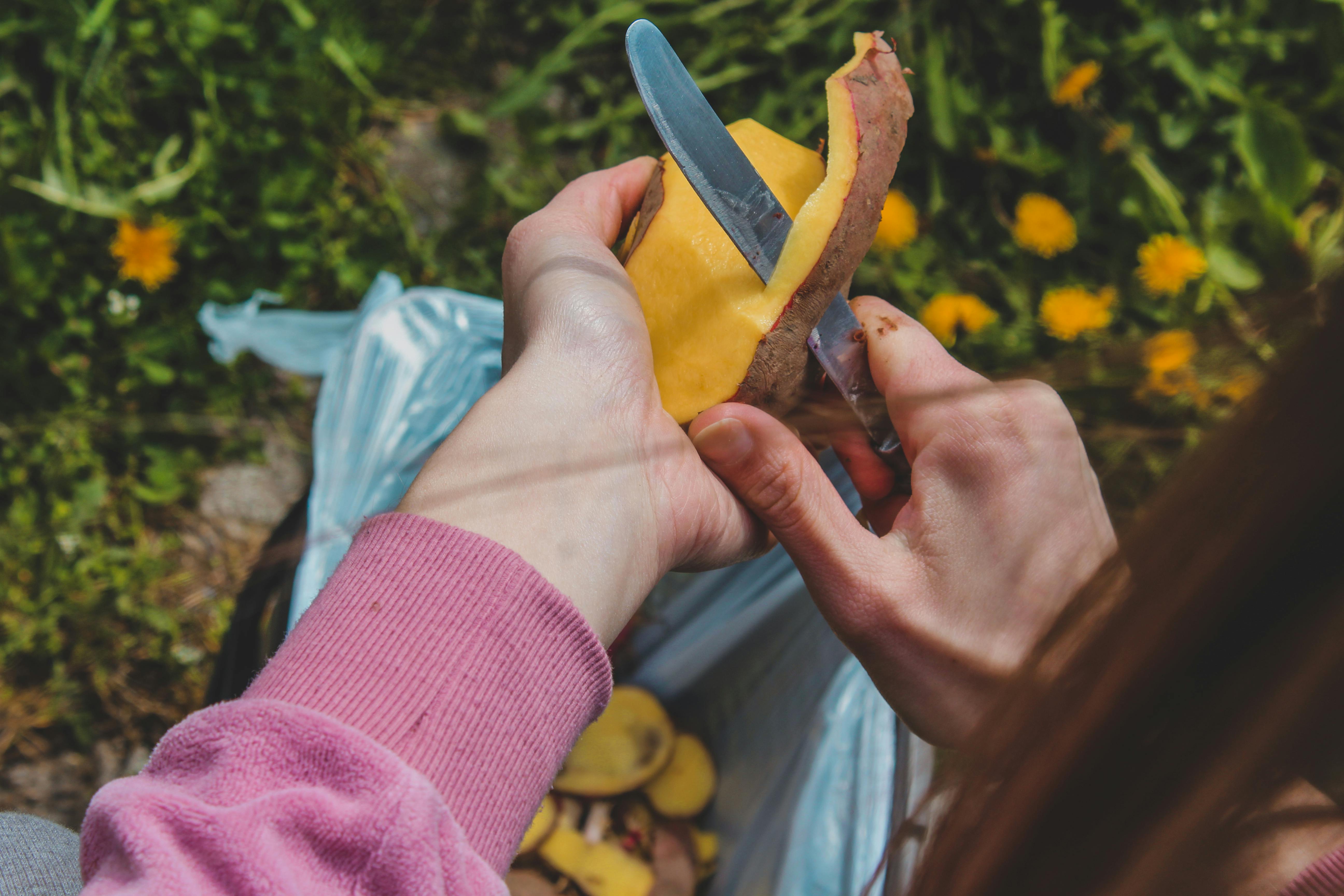 Hands peeling potatoes with a knife outdoors, showcasing food preparation and fresh produce.