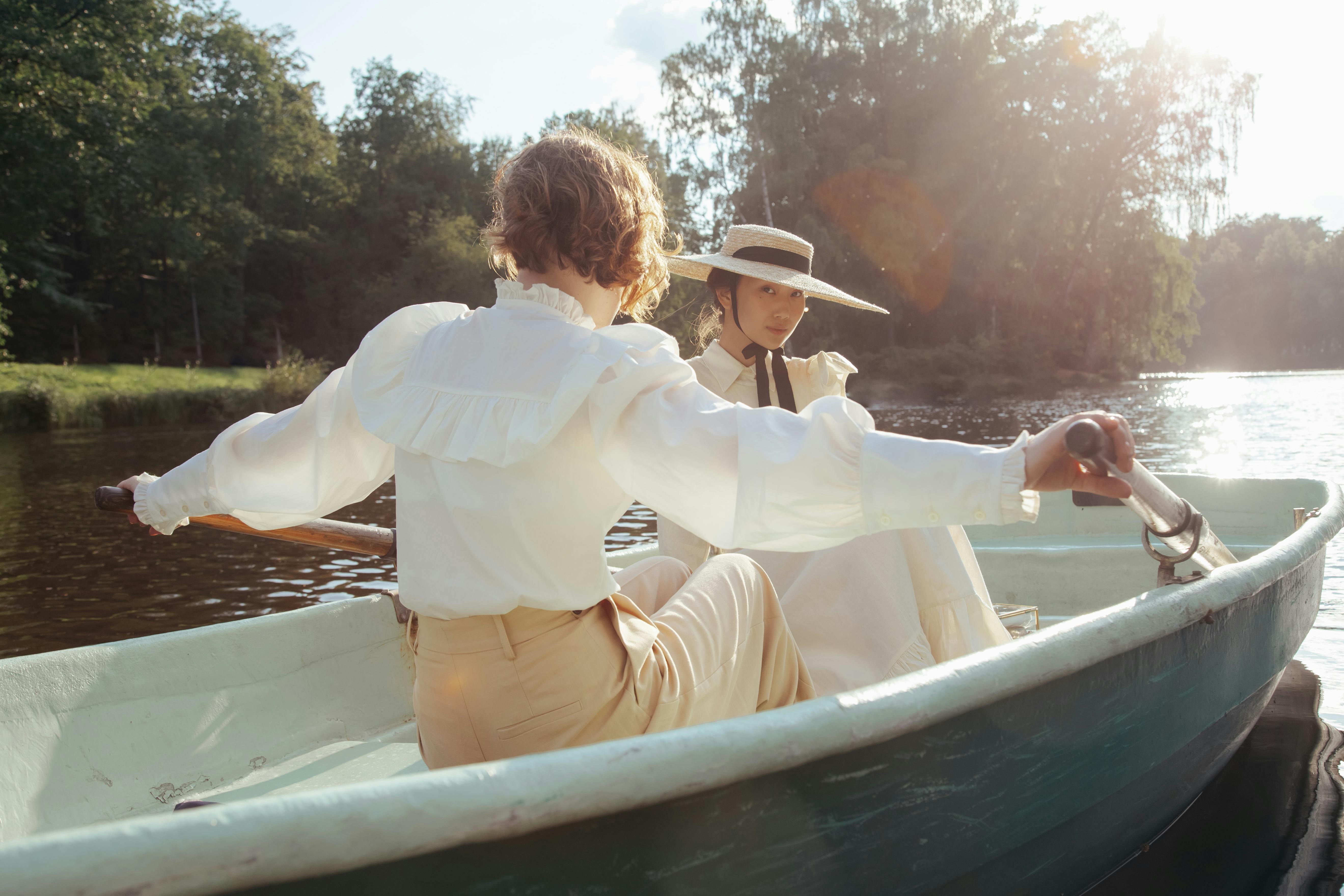 Two women in vintage attire enjoying a peaceful rowing experience on a tranquil lake.