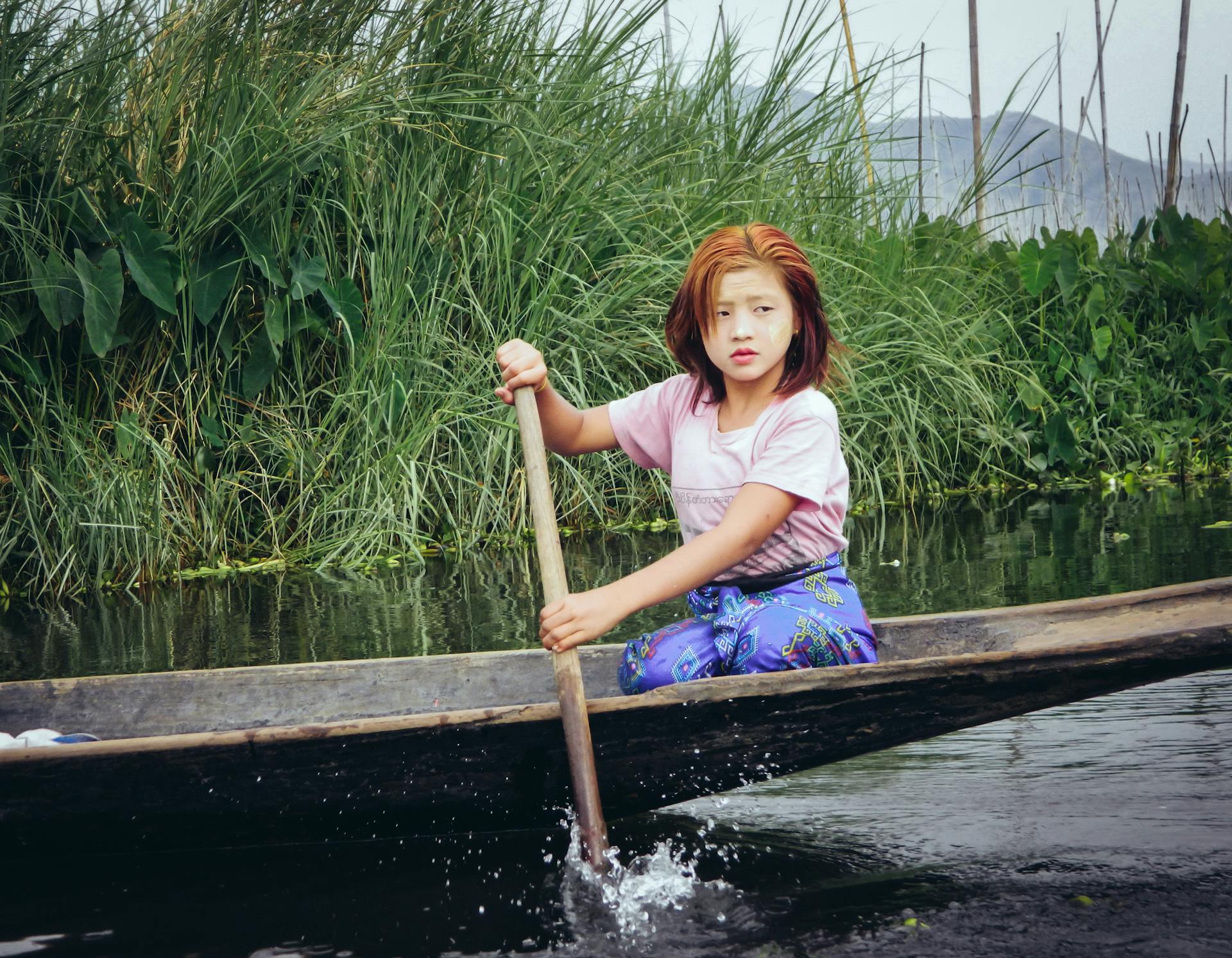 Young girl paddling a wooden boat on a lush waterway in Shan, Myanmar, showcasing local culture.