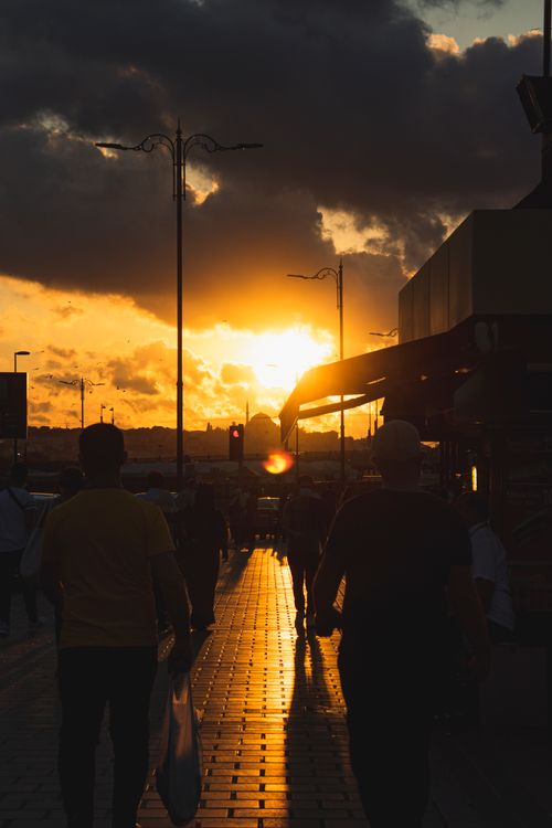 Free Silhouetted pedestrians walk under a vibrant sunset sky on a lively city street. Stock Photo