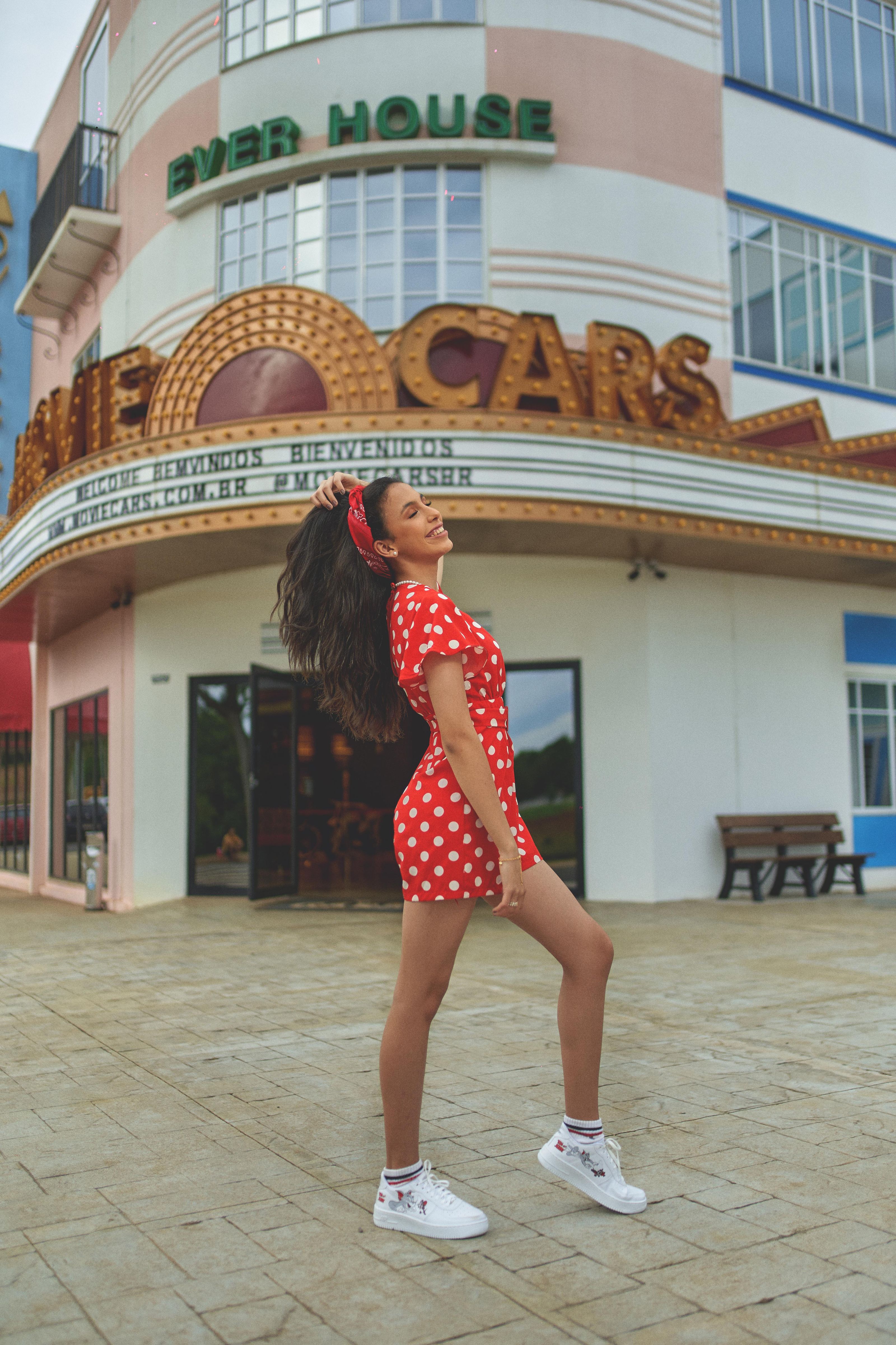 Jovem mulher em um vestido vermelho com poás brancos posa confiante do lado de fora de um teatro vintage.