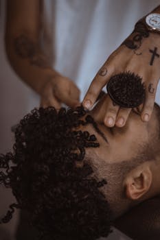 Close-up of a barber styling a man's curly hair, showcasing detailed hand tattoos and grooming tools.
