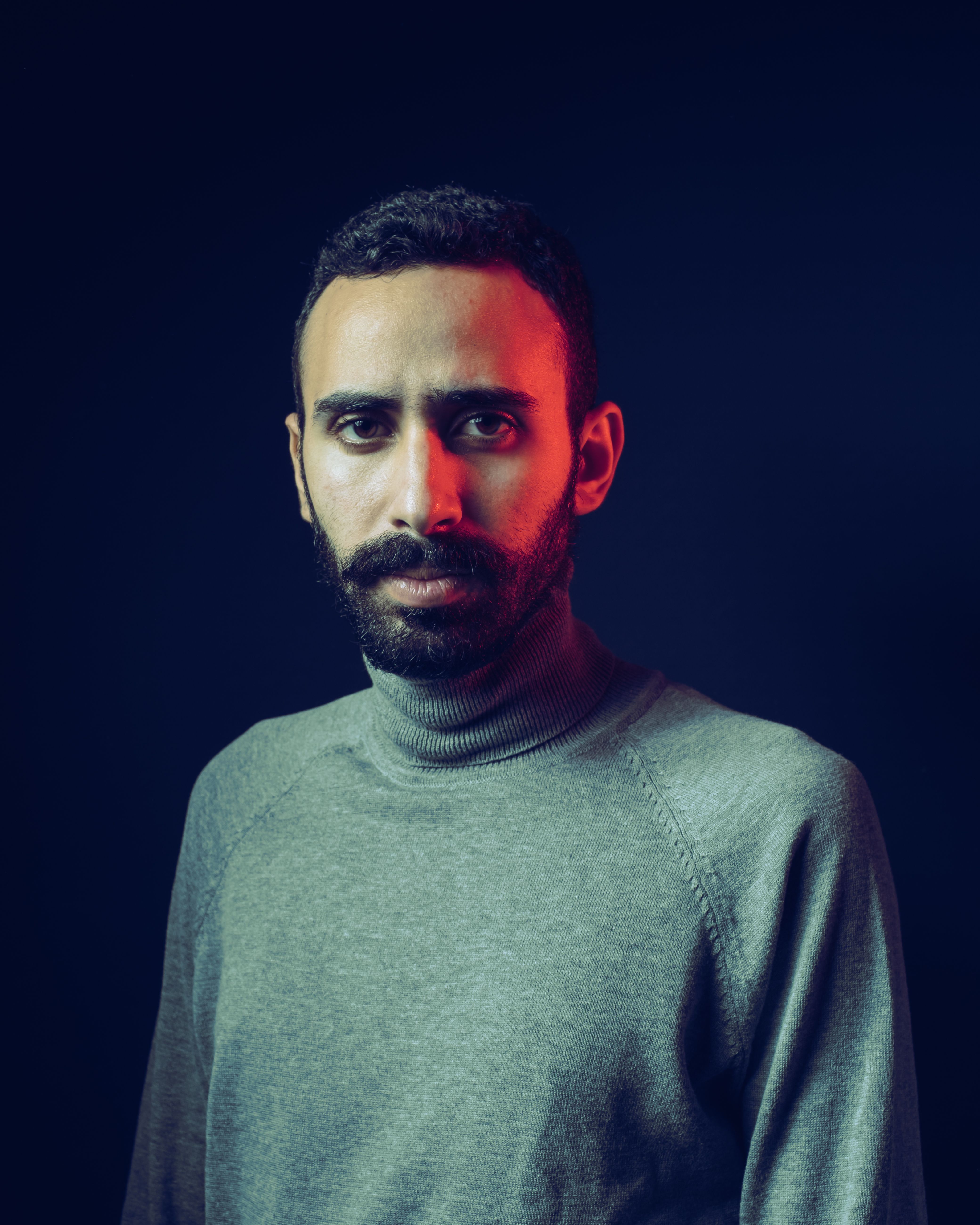 A serious bearded man in a sweater posed against a dark background in a studio