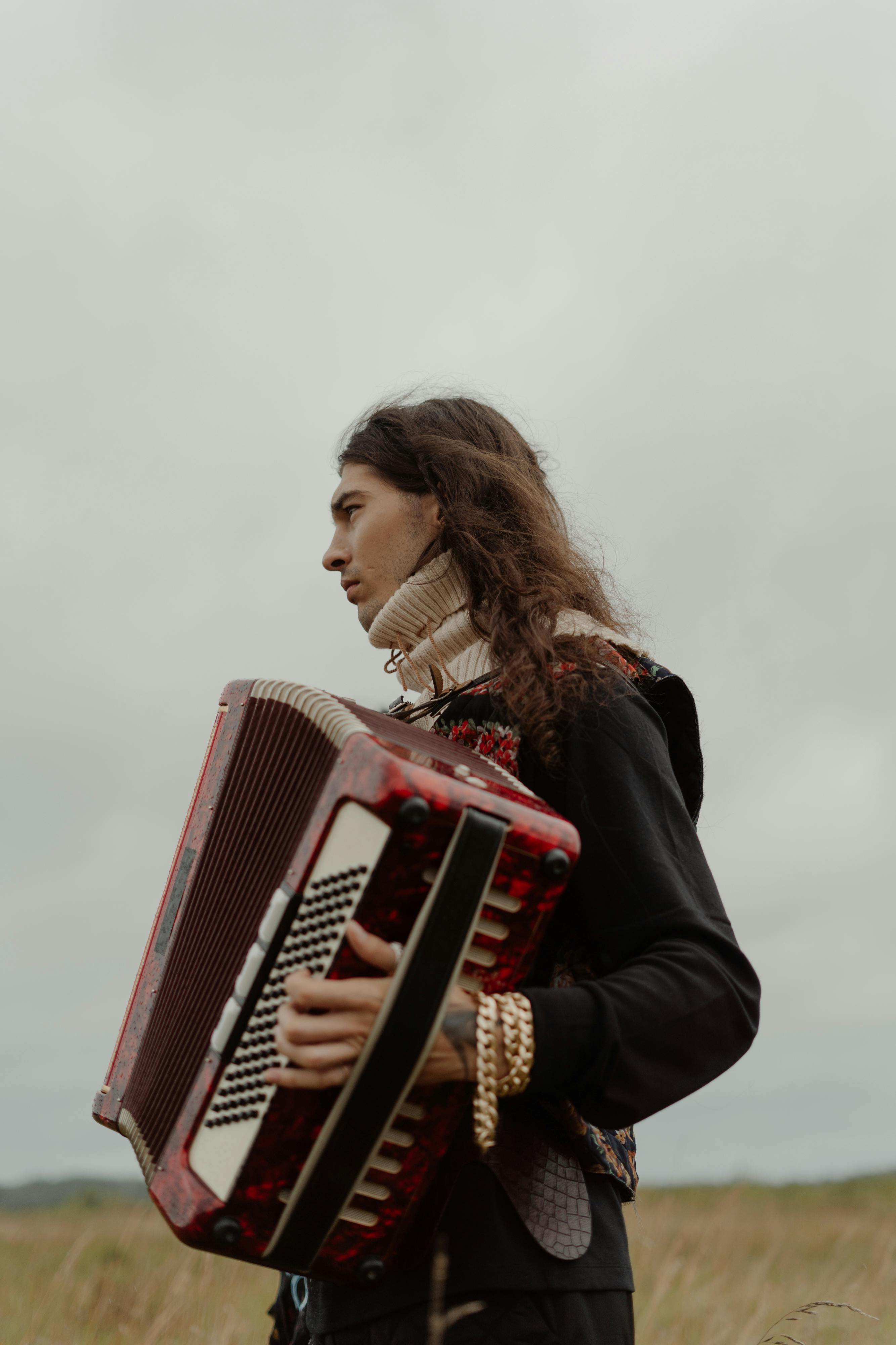 Portrait of a young musician playing an accordion in a field under cloudy skies.