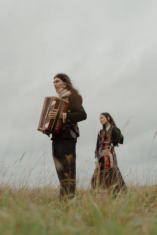 Man Playing Accordion on Grassland with Woman in Dress Standing behind