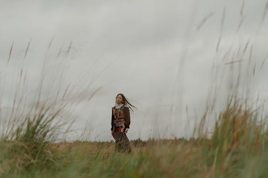A bohemian woman walks through a grassy field under a cloudy sky, exuding a sense of freedom and style.
