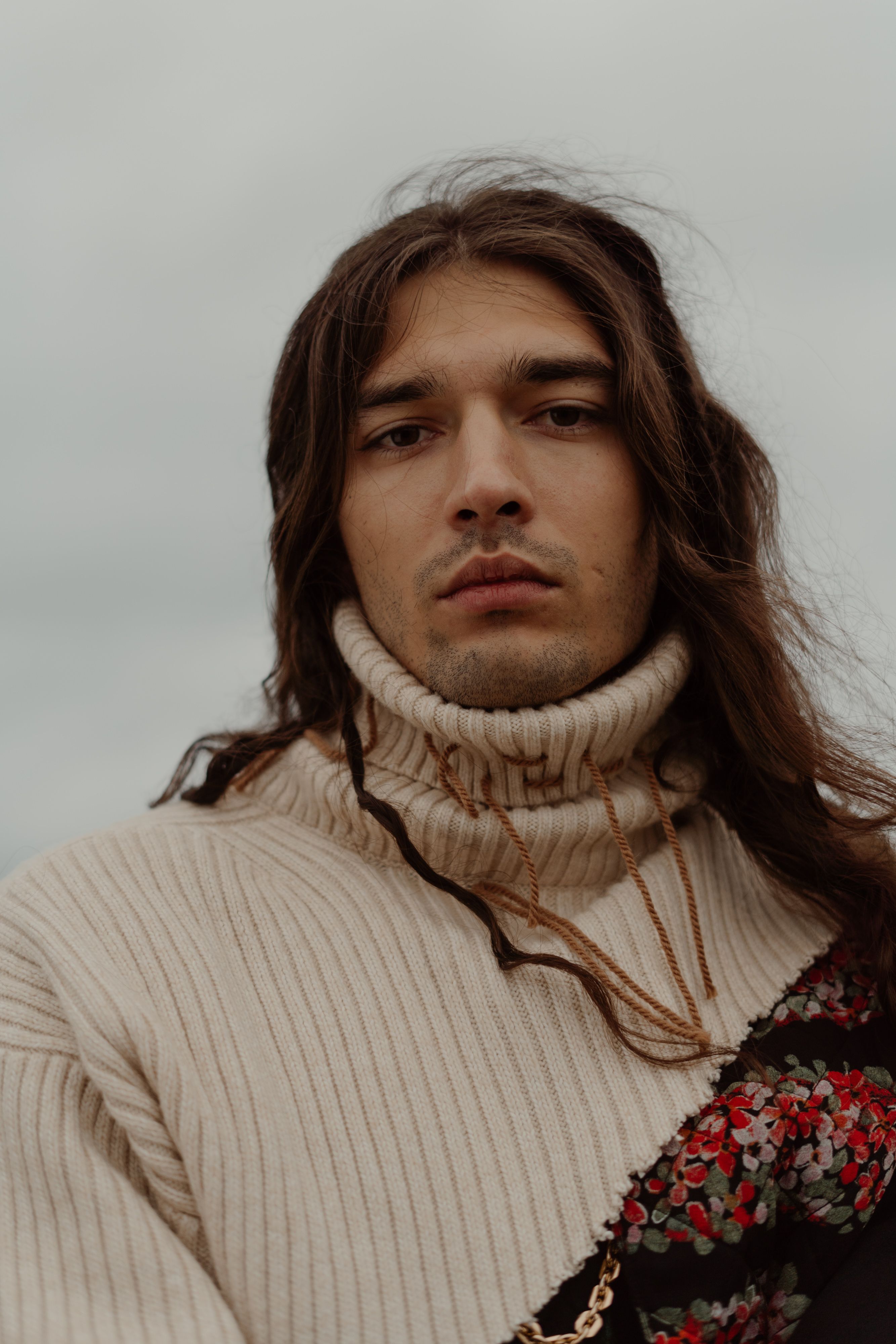 Close-up portrait of a young man with long hair in a knitted sweater outdoors.