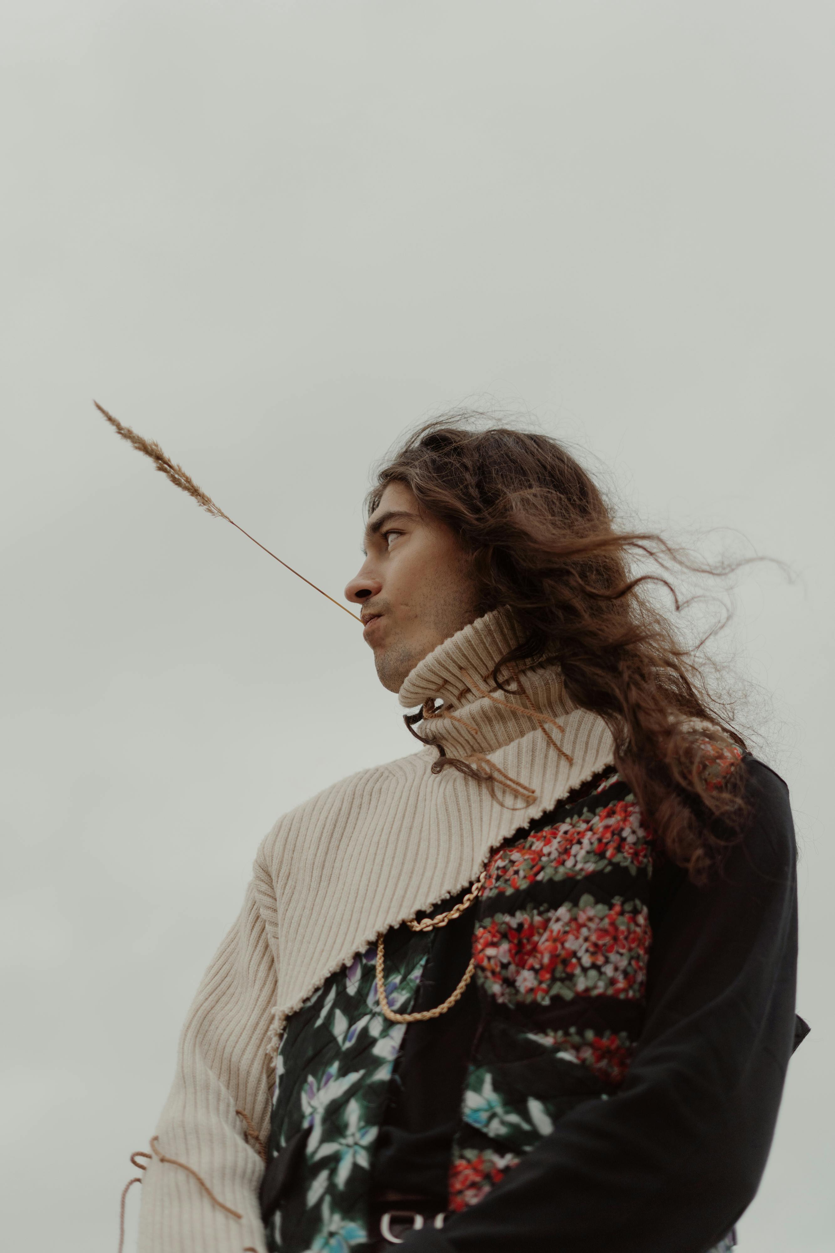 Artistic portrait of a young man with long hair wearing a textured sweater holding straw in mouth outdoors.