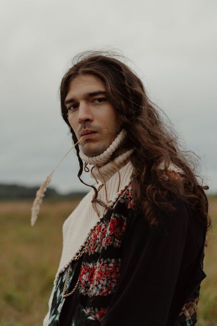 Portrait of a young man with long hair and sweater, set in a serene outdoor field.