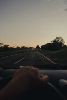 A hand on the steering wheel driving on a rural road during sunset, capturing the serene twilight scene.