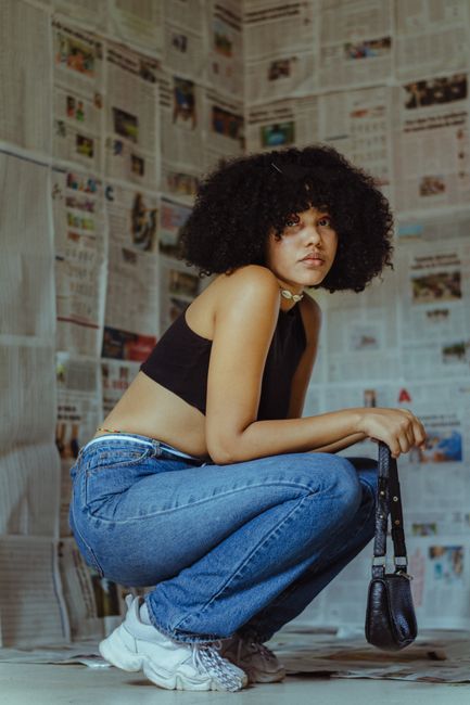 A young woman with curly hair and casual outfit crouching in a newspaper-covered room.