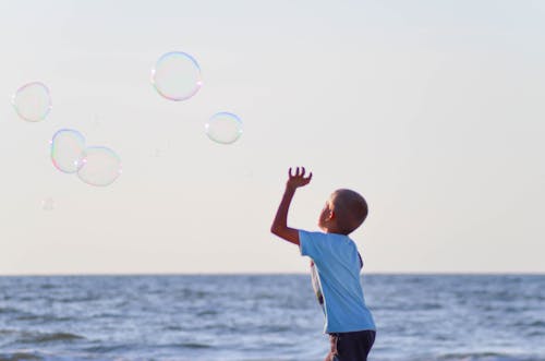 Ragazzo In Maglietta Bianca Che Gioca Bolle Vicino Al Corpo D'acqua Sotto Il Cielo Grigio Durante Il Giorno