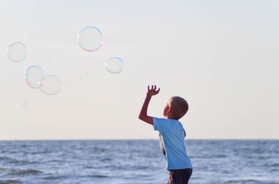 Free Boy in White T Shirt Playing Bubbles Near Body of Water Under Grey Sky during Daytime Stock Photo