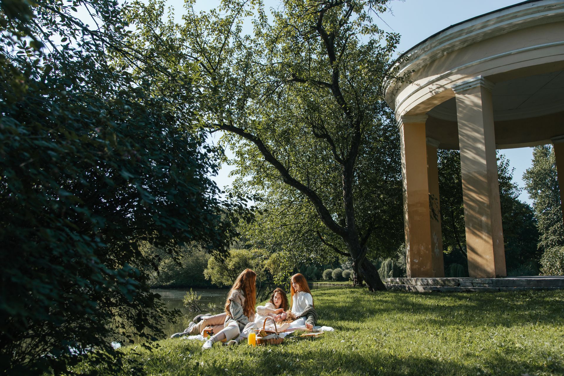 Three women enjoy a serene picnic near a historical pavilion in a lush park.