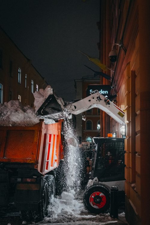 Free An excavator loads snow into a truck on a city street during nighttime winter clearing. Stock Photo
