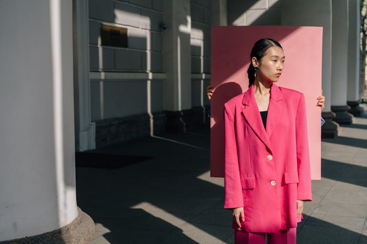 Person Holding A Pink Sheet Behind A Woman In Pink Blazer