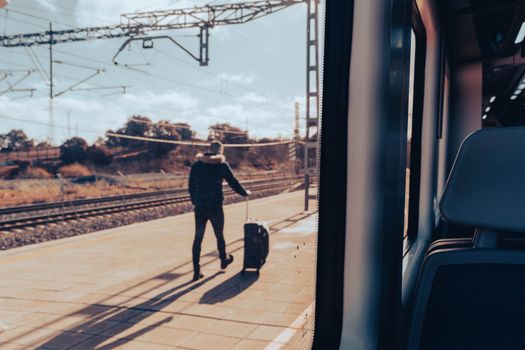 A man walks on a train platform with luggage, viewed from a train window.