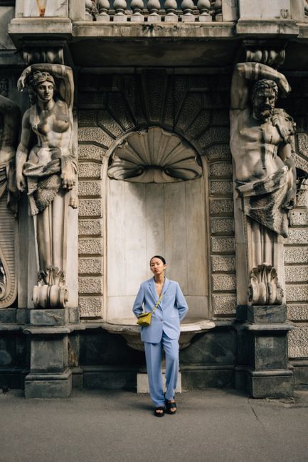 Woman in blue suit stands elegantly by historical statues in urban setting.