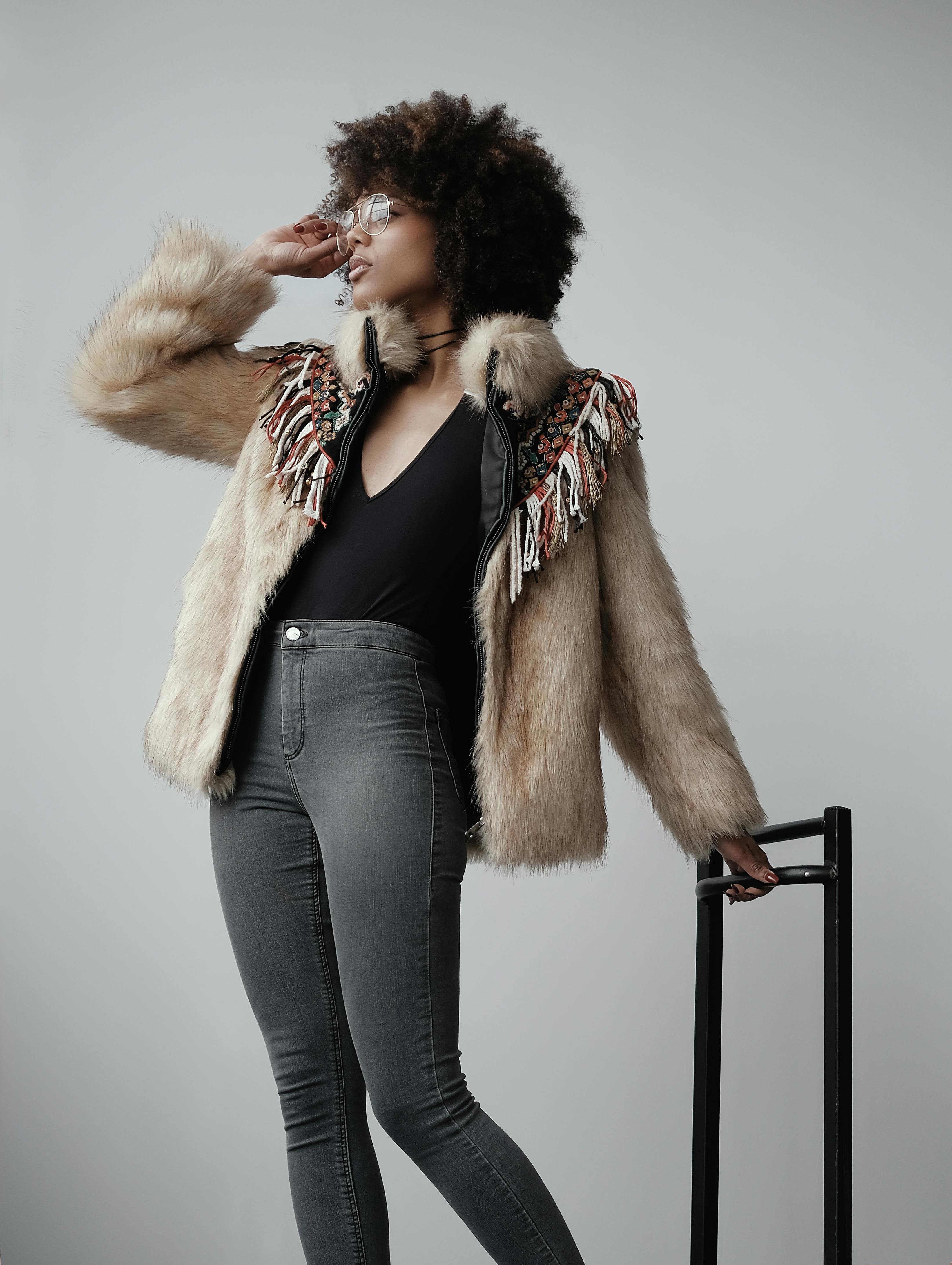 Chic woman in fur coat and glasses poses with luggage against gray background.