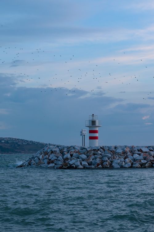 Free Lighthouse on rocky shore with calm water and blue sky in the background. Stock Photo