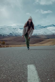 A woman in winter clothing stands on a road with snow-capped mountains in the background. Low angle shot.