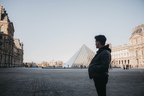 Free A man stands in Louvre courtyard, gazing at the iconic glass pyramid in Paris. Stock Photo