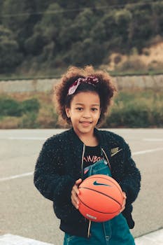 African American girl smiling and holding a basketball outdoors in a casual setting.