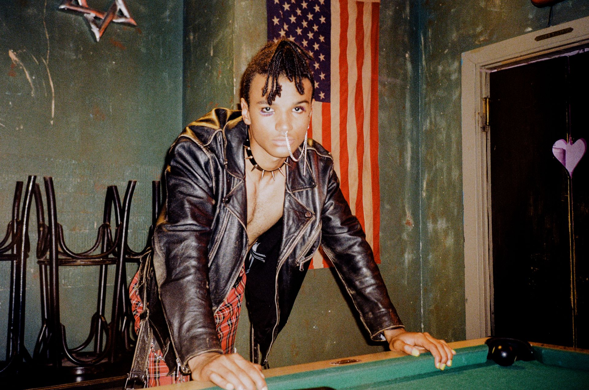 A confident person in a leather jacket leaning on a pool table indoors with an American flag backdrop.