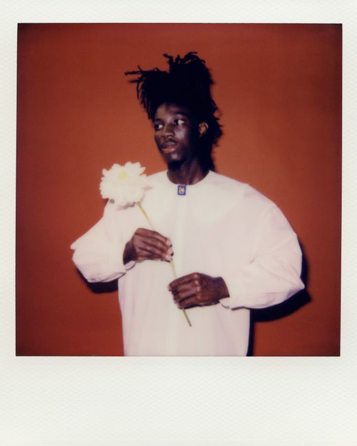 A stylish man with braided hair holds a white flower against a warm background in this portrait.
