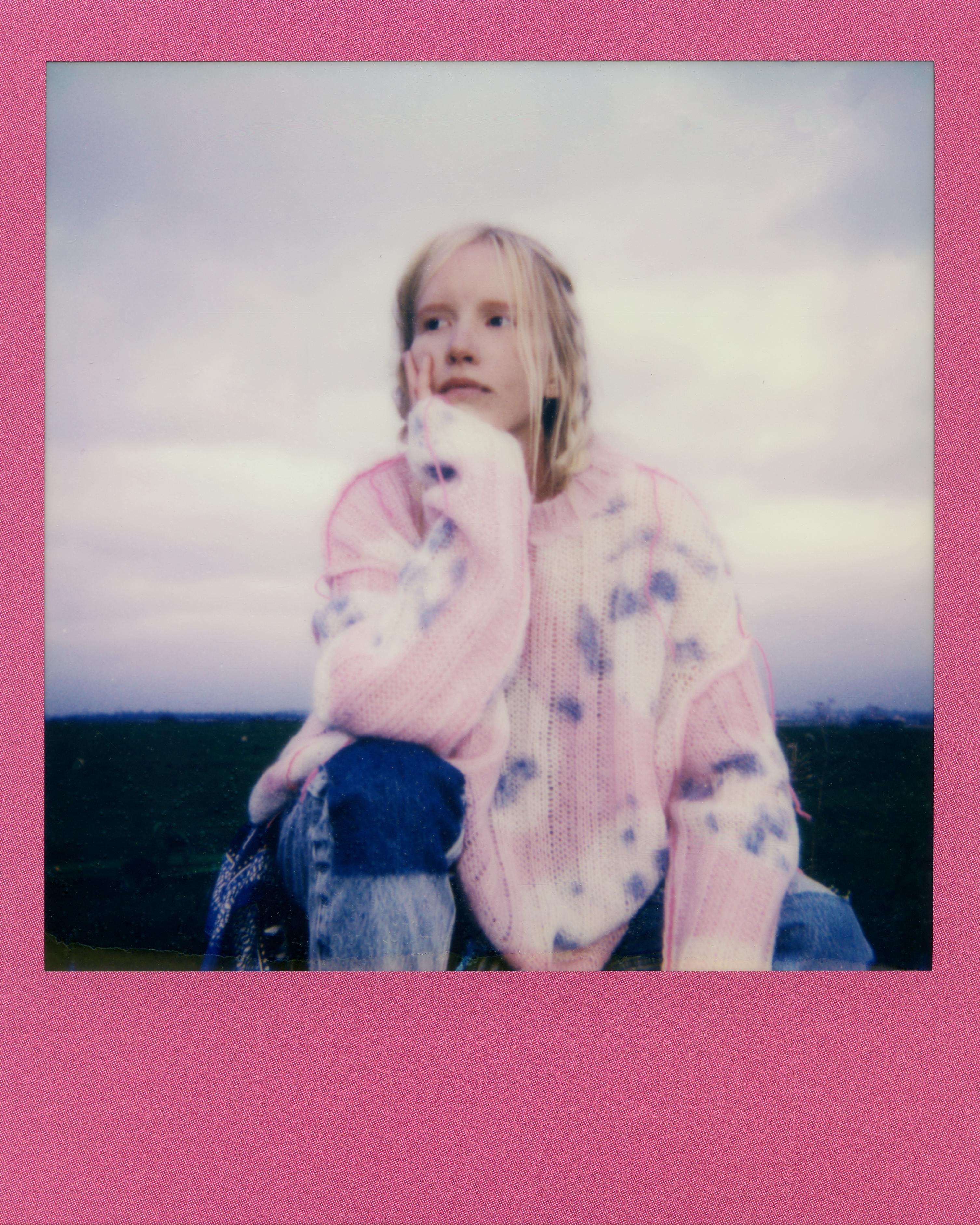 A young woman in a pink tie-dye sweater sitting outdoors with a thoughtful pose.