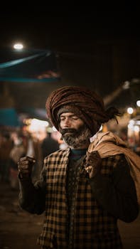 An elder man in traditional clothing stands in a busy New Delhi market at night.