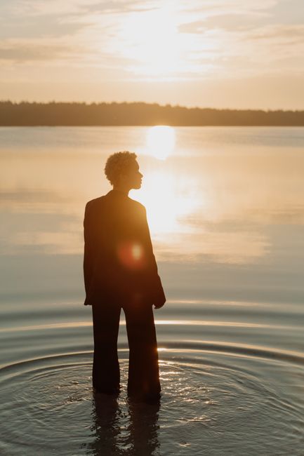A woman in a suit stands in water at sunset, creating a serene silhouette with ripples.